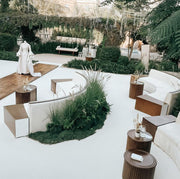 Modern outdoor seating area with white sofas, wooden tables, and greenery.