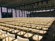 Rows of white chairs with gold frames in a large indoor venue. Moments rentals