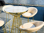 Gold-framed table with marble top surrounded by beige chairs on a dock.
