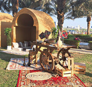 Outdoor setting with a wooden wagon, cushions, and decorative items on a grassy area with palm trees.