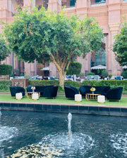 Outdoor seating area with black chairs and tables by a poolside, with a building and trees in the background.