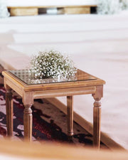 Wooden side table with a glass top featuring baby's breath flowers, placed on a bed.