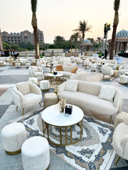 Outdoor seating area with beige sofas, ottomans, and coffee tables on a patterned rug with palm trees and buildings in the background.