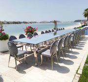 Long outdoor dining table with chairs by a waterfront, featuring decorative flowers.
