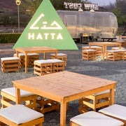 Outdoor seating area with wooden tables and stools in front of a green triangular sign with 'HATTA' branding.