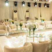 Elegant event setup with beige sofas, glass tables, and floral arrangements in a large room.