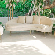 Beige curved sofa with gold legs in an outdoor setting with greenery and flowers.