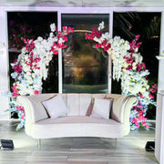 Decorative floral arch with pink and white flowers in front of a window with a sofa underneath.