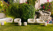 Outdoor setting with white chairs and ottomans on a grassy area with a fountain and garden in the background.