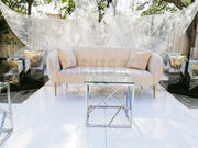 Beige sofa with cushions and a glass coffee table on a white floor with transparent plastic sheeting in the background.