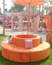 Orange circular couch with pink cushions on a grassy area with palm trees and colorful decorations in the background.