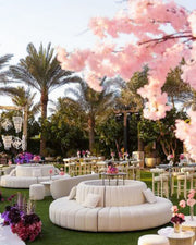 Outdoor event setup with white round couches, tables, and pink floral decorations in a garden setting.