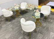 Modern white chairs with gold legs arranged around small round tables with floral arrangements in a room.