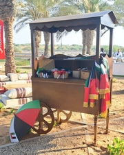 Wooden cart with a colorful umbrella and various items on wheels, set against a palm tree-lined background.