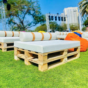 Wooden pallet furniture with white cushions on a grassy area with buildings in the background - Moments Rentals