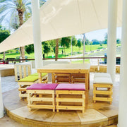 Outdoor seating area with wooden tables and benches under a large white canopy, surrounded by greenery.