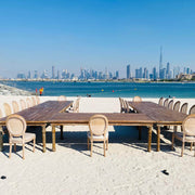 Outdoor dining setup with tables and chairs on a sandy beach, city skyline in the background.
