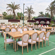 Outdoor setting with wooden tables and chairs arranged for an event, palm trees in the background.